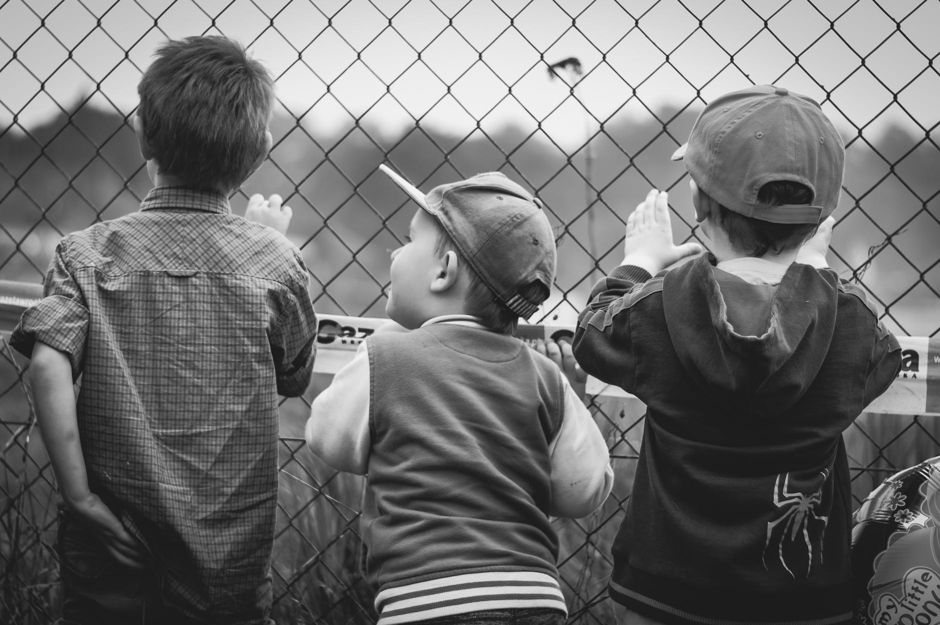 grayscale photography of three boys facing towards fence